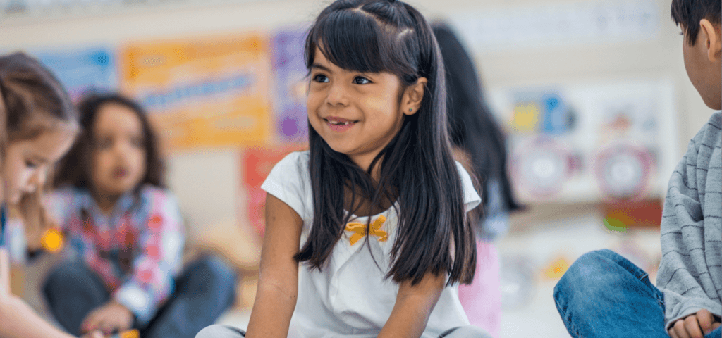 A smiling young girl sits on the floor in a classroom with other children blurred in the background.