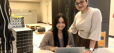Two researchers stand in a data centre beside server racks while one works on a laptop, highlighting collaboration in high-performance computing and data infrastructure.