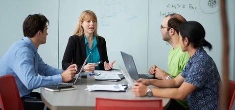 A group of researchers and students sit around a table in discussion, laptops open, collaborating on ideas in a classroom or research meeting setting.