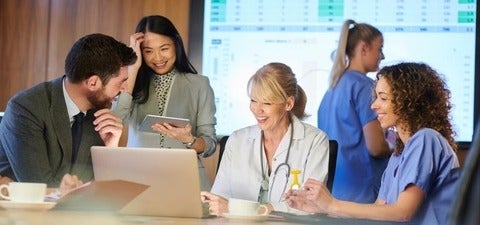 A multidisciplinary healthcare team gathers around a laptop, smiling and reviewing data during a collaborative meeting, with digital dashboards displayed in the background.