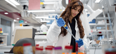A scientist in a lab coat and blue gloves uses a pipette while working at a laboratory bench.