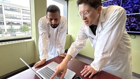 Two researchers in lab coats review information on a laptop near a window, with data visualizations displayed on a screen behind them.
