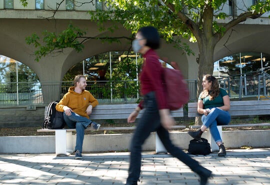 Students sitting on campus bench as masked student walks past