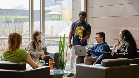Four students sit in a lounge, one stands and gestures to a laptop