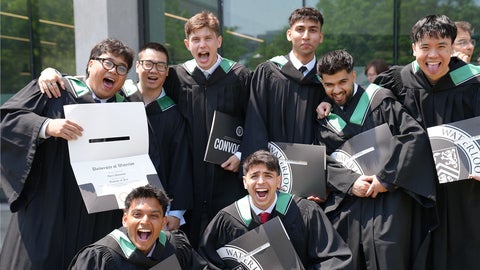A group of Arts graduates in convocation gowns shout and hold up their diplomas