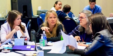 Four women in discussion at a table with papers and supplies