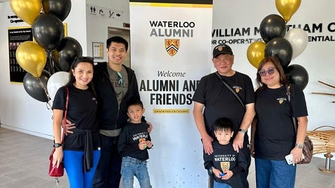 A set of grandchildren, parents, and grandparents stand in front of a UW alumni sign