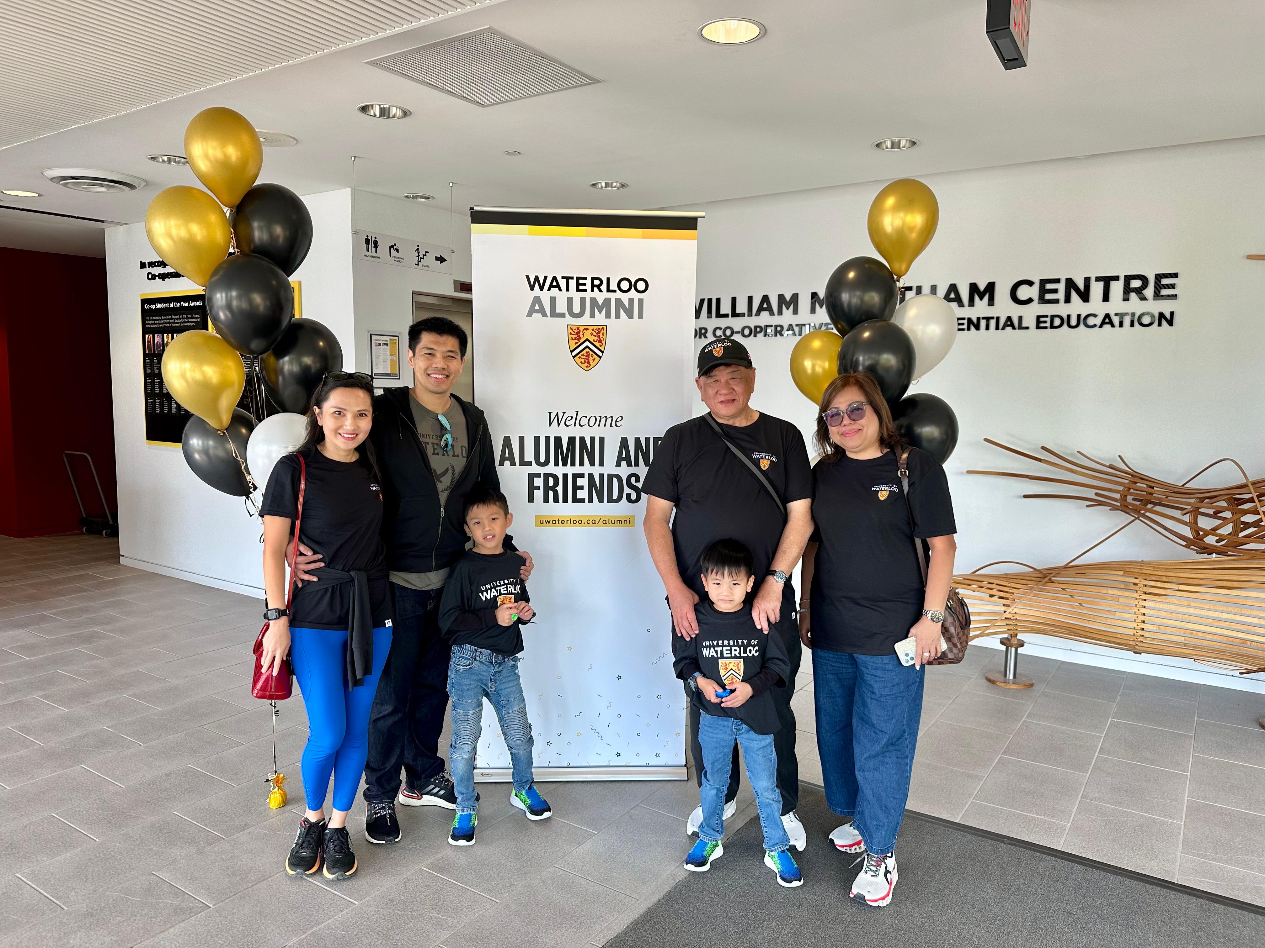 Grandparents, parents, and children of the Tee family in front of an alumni banner