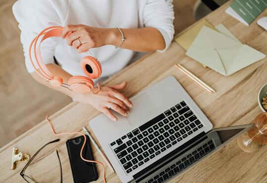 person holding orange headphones while sitting in front of a laptop with finger on the trackpad