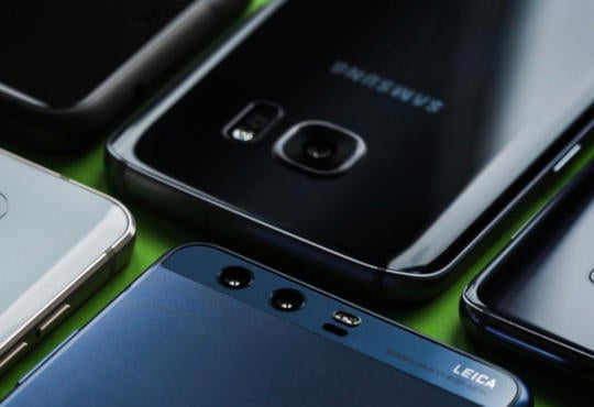 A photo of six smartphones of various brands lying face down in two rows on a green surface