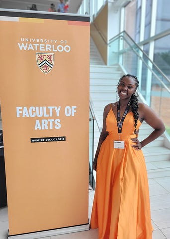 Graduate Emily Radliffe, dressed in a flowing orange gown, stands next to the Faculty of Arts banner