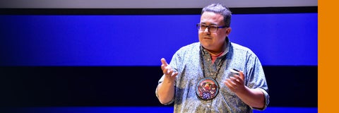Jay Havens at a speaking event. He is wearing a medalian around his neck of intricate Indigenous beadwork