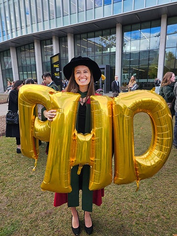 A PhD grad in their red and green robes holds up giant gold balloons that spell out P H D