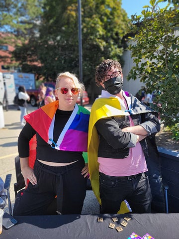 Two people post at an event, one is wraped in rainbow Pride flag, the other has their arms crossed in a confident manner