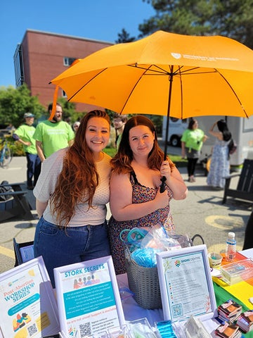 two students underneath a big orange umbrella standing at a table with pamphlets