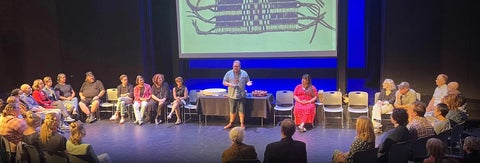 Jay Havens speaks to a group encircling the stage, there are Indigenous items including a wampum belt, on display