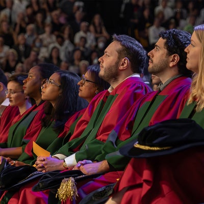 Arts PhD graduates site in a row at convocatin, all wearing their red and greedn robes and holding their black hats