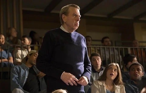 Film still of a middle aged man standing up to speak amongst students in a lecture hall
