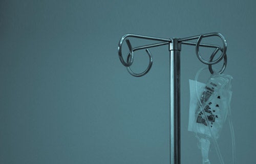 An intravenous fluids bag hangs on an IV pole. The image is tinted teal to imply a sense of foreboding