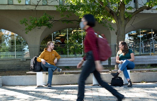 Two students sit at oppostie ends of a bench outside Dana Porter Library while a student wearing a mask walks by