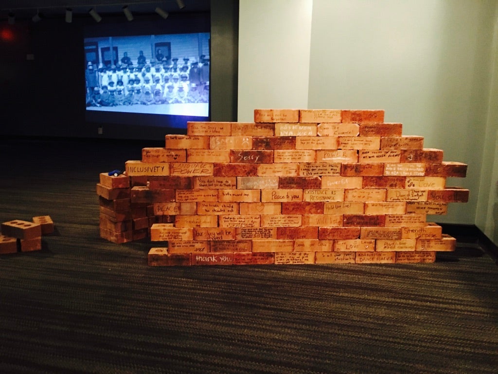 Detail of a gallery exhibit showing a small brick wall, and each brick has a name on it