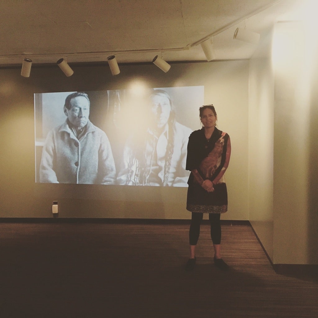 A woman stands next to a projected image of two Indigenous People, she is giving a tour or a talk