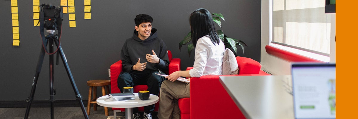 A student works with a faculty member in a user experience lab. There are post notes in the background showing a UX exercise, a camera on a tripod, and a laptop in the foreground