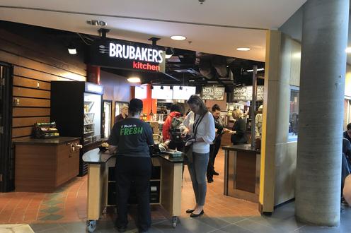 A woman reaching into her purse to pay for food at a register. The sign above reads BRUBAKERS kitchen.