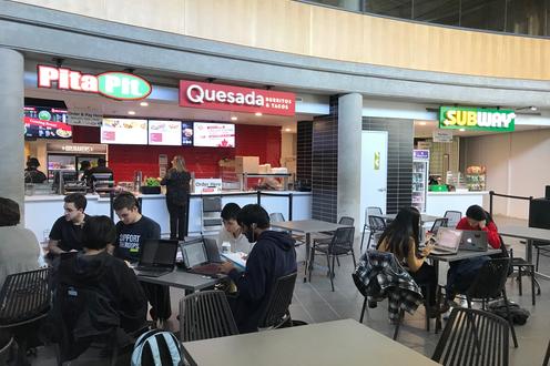 Students in foreground at tables in the updated dining space in the SLC with Pita Pit, Quesada, and Subway in the background.