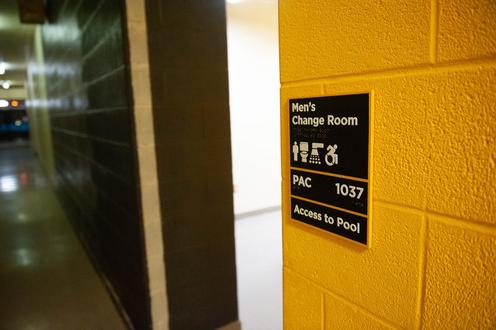 Black sign on yellow brick wall with text: Men's Change Room PAC 1037 Access to Pool. Sign includes braille and icons.