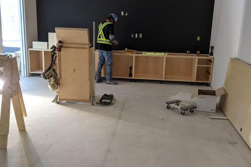 A millworker in the background installing cabinetry in a room under construction.