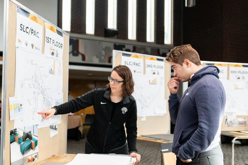 A woman in black sweater points to a board with floor plans as male student looks on thoughtfully.
