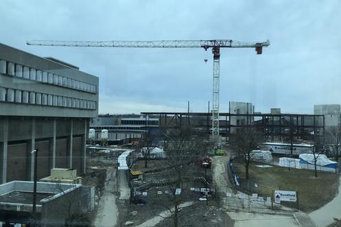 Large white crane over a construction site with steel beams outlining a new building and existing MC building on the left.