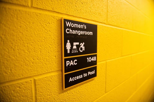 Gold wall with black sign with the words: Women's Changeroom PAC 1048 Access to Pool. Icons for women, showers, and accessible