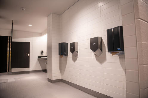 Paper towel and hand dryers on a freshly tiled white wall with sink to the far left.