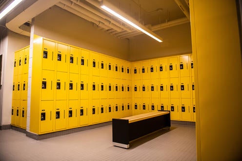 A large room with yellow lockers 3 rows high on all three walls and bench in the middle