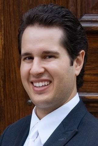 Photo of a man with black hair smiling at the camera. Wearing black suit jacket and white collared shirt along with a white tie.