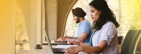 Horizontal oriented web banner of two students at desks working individually on their laptops. Light yellow border framing the banner. 