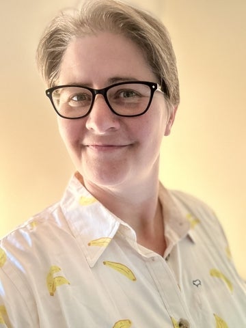 photograph of person with dark coloured rectangular glasses, smiling at the camera, wearing a white button down shirt with images of bananas on it. 