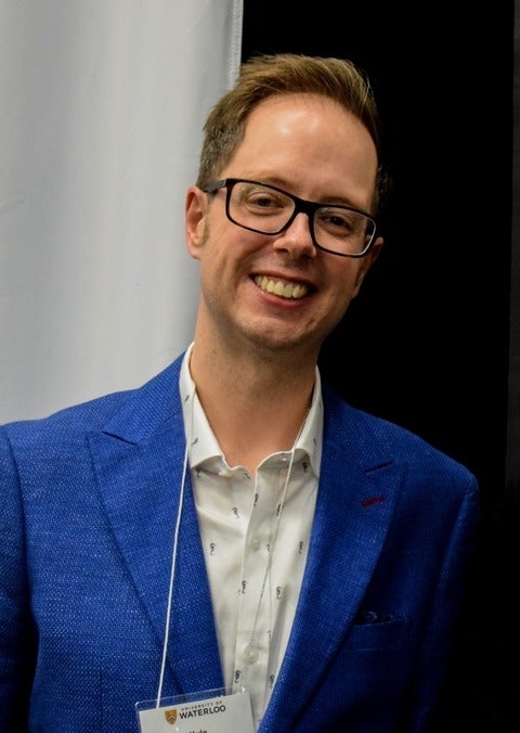 headshot of a person smiling with short hair, glasses, white shirt and blue blazer
