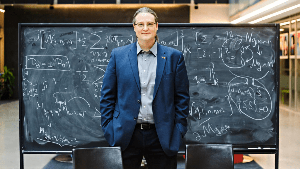 Dr. Avery Broderick standing in front of a blackboard covered with mathematics
