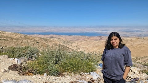 Aparajita, a graduate student standing in an open area with sand dunes, plants, and a source of water in the background