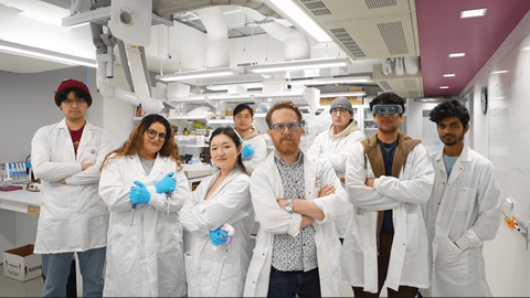 Eight scientists in the NeurdyPhagy lab. They are all posing with their arms crossed. The scientists are wearing white lab coats with blue gloves.