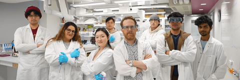 Link, Eight scientists in the NeurdyPhagy lab. They are all posing with their arms crossed. The scientists are wearing white lab coats with blue gloves.