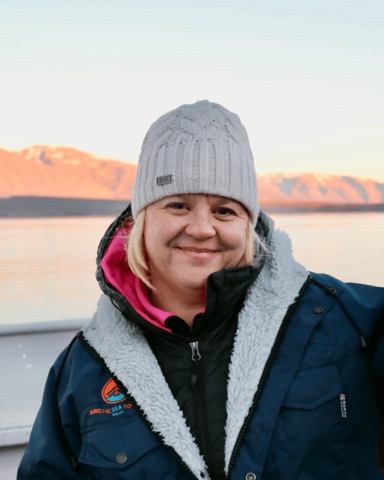 A headshot of Kirsten. She is wearing a grey winter hat and a blue winter coat. There is a lake and white capped mountains in the background. 