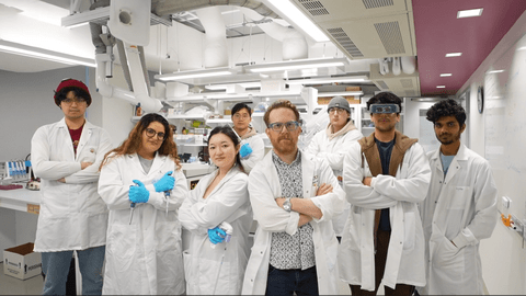 Eight scientists in the NeurdyPhagy lab. They are all posing with their arms crossed. The scientists are wearing white lab coats with blue gloves.