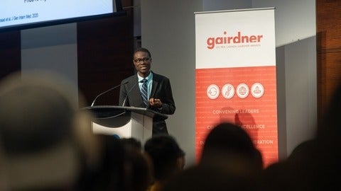 Dr. Abiola Olaitan speaking in front of a crowd. There is a pull up banner behind him that says "gairdner" in larger letters and "LES PRIX CANADA GAIRDNER AWARDS" in smaller letters. 
