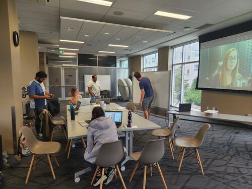 Students collaborating in class and virtually. Four students sitting around a table and one person drawing on a white board. One virtual student is speaking while her image is projected on another screen.