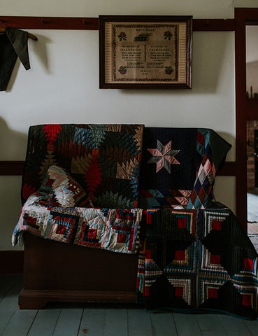 a photo of two handed quilted blankets draped over a bench, against a white wall, hanging above is an old framed piece of fraktur text art.