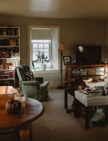 A photo of Brubacher's apartment living room. Featuring a deep silled window, book shelves full of games and colourful books, a dining table, and a comfy green chair. 